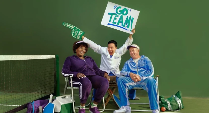 Three people sitting near a tennis net. One person is holding a 'Go Team' sign, dressed in a dentist's coat, and raising their arms.